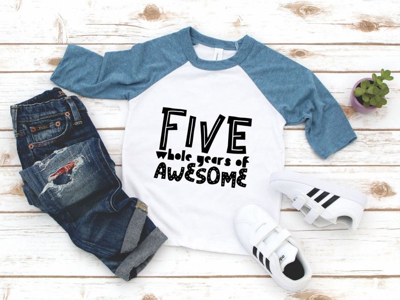 May include: A white and blue baseball tee shirt with the text "FIVE whole years of AWESOME" in black. The shirt is on a white wooden surface with a pair of white trainers and blue jeans.