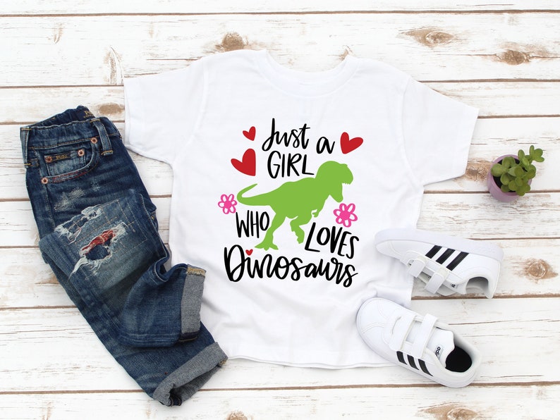 Może przedstawiać: Biała koszulka z zielonym dinozaurem i napisem "Just a girl who loves dinosaurs". Koszulka leży na białym drewnianym tle z niebieskimi dżinsami i białymi butami.