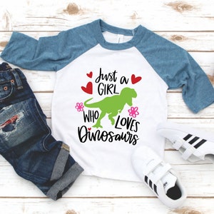 Może przedstawiać: Biało-niebieska koszulka typu raglan z napisem "Just a girl who loves dinosaurs" i zielonym dinozaurem. Koszulka leży na białym drewnianym tle z parą białych but&oacute;w sportowych i niebieskimi dżinsami.