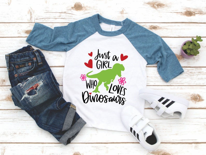 Może przedstawiać: Biało-niebieska koszulka typu raglan z napisem "Just a girl who loves dinosaurs" i zielonym dinozaurem. Koszulka leży na białym drewnianym tle z parą białych but&oacute;w sportowych i niebieskimi dżinsami.