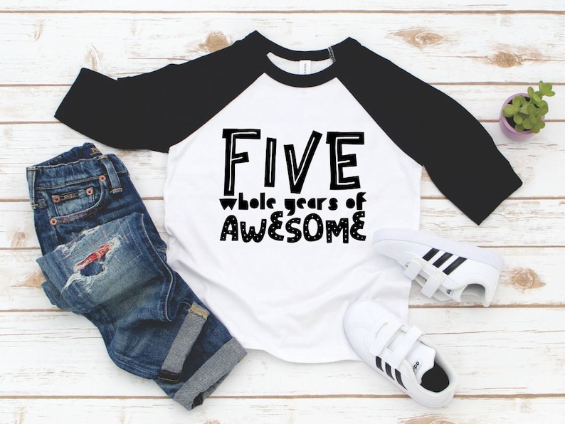 May include: White and black baseball tee shirt with the text "FIVE whole years of AWESOME" in black. The shirt is on a white wooden surface with a pair of white trainers and blue jeans.