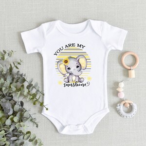 May include: White baby onesie with a gray and yellow striped background. A cartoon elephant with a sunflower on its head is sitting on the stripes. The text "You are my sunshine" is printed below the elephant.