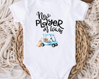 May include: White baby onesie with the text "New Player in Town" in black script. A watercolor illustration of a blue golf cart with golf clubs is below the text. The onesie is displayed on a woven basket.