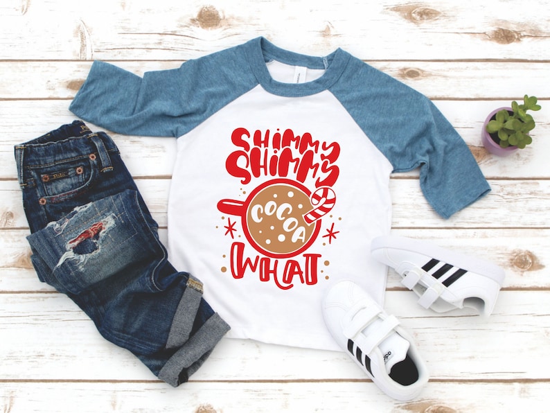 May include: A white and blue baseball tee shirt with red text that says "Shimmy Shimmy Cocoa What". The shirt features a brown mug with a candy cane and a white background.