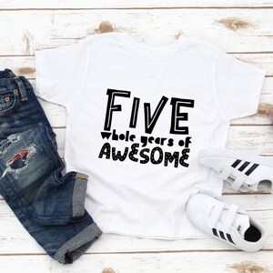 May include: White t-shirt with black text that reads "FIVE whole years of AWESOME". The shirt is laid out on a white wooden surface with a pair of white trainers and blue jeans.