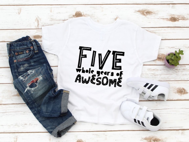 May include: White t-shirt with black text that reads "FIVE whole years of AWESOME". The shirt is laid out on a white wooden surface with a pair of white trainers and blue jeans.