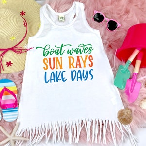 May include: White tank top with a frayed hem, featuring the text "boat waves sun rays lake days" in colorful letters. The tank top is on a pink fuzzy surface with a straw hat, sunglasses, and beach toys.
