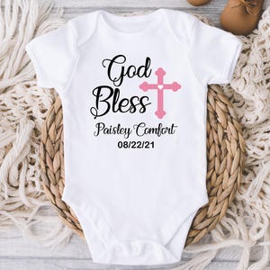 K&ouml;nnte beinhalten: Wei&szlig;er Baby-Body mit dem Text "God Bless" und einem rosa Kreuz. Der Body enth&auml;lt auch den Namen "Paisley Comfort" und das Datum "08/22/21". Der Body wird auf einem geflochtenen Korb pr&auml;sentiert.