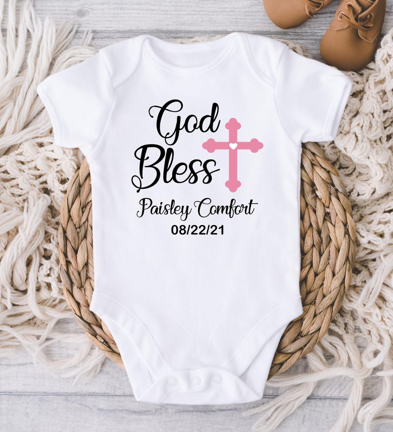 K&ouml;nnte beinhalten: Wei&szlig;er Baby-Body mit dem Text "God Bless" und einem rosa Kreuz. Der Body enth&auml;lt auch den Namen "Paisley Comfort" und das Datum "08/22/21". Der Body wird auf einem geflochtenen Korb pr&auml;sentiert.