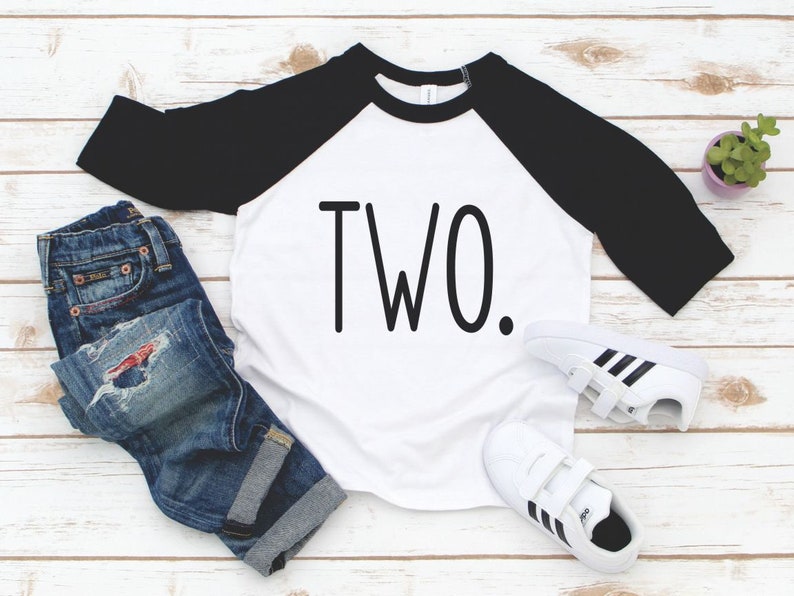 May include: White and black baseball tee shirt with the word "TWO" printed in black on the front. The shirt is on a white wooden surface with a pair of blue jeans and white trainers.