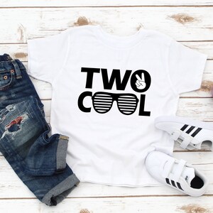 Może przedstawiać: Biała koszulka z czarnym napisem "TWO COOL" z okularami przeciwsłonecznymi i znakiem pokoju. Koszulka leży na białym drewnianym tle z parą białych but&oacute;w sportowych z czarnymi paskami i małą doniczką z rośliną.