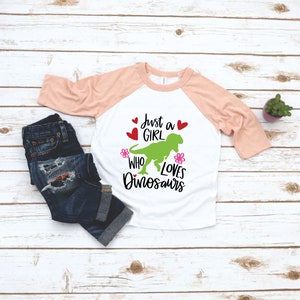 Może przedstawiać: Biało-brzoskwiniowy t-shirt z zielonym dinozaurem i napisem "Just a girl who loves dinosaurs". Tekst jest czarny z r&oacute;żowymi kwiatami i czerwonymi sercami.