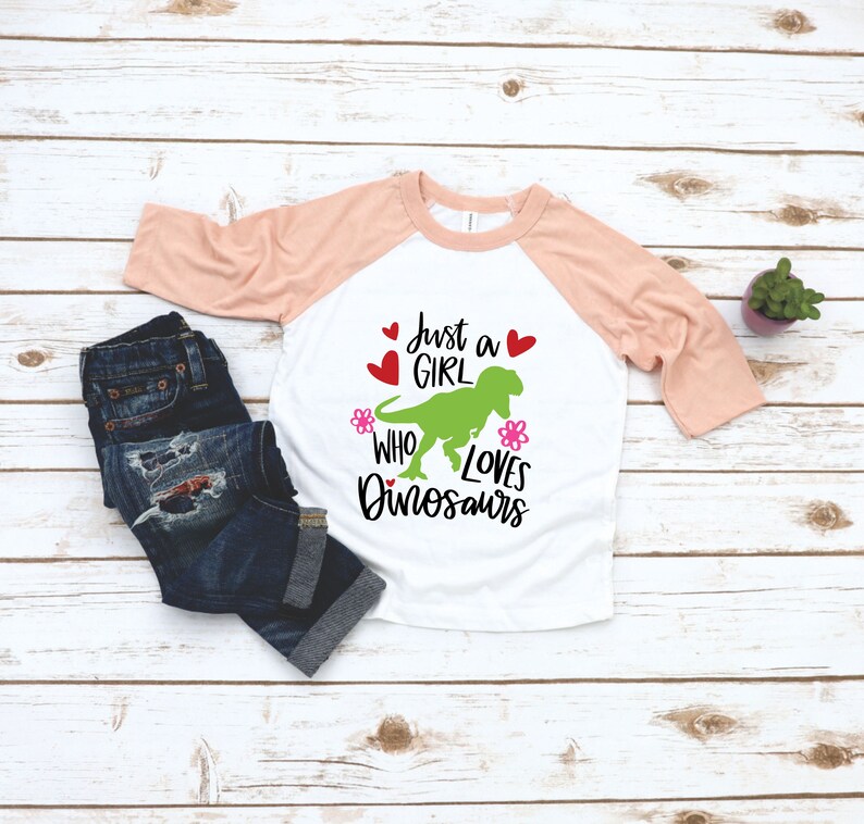 Może przedstawiać: Biało-brzoskwiniowy t-shirt z zielonym dinozaurem i napisem "Just a girl who loves dinosaurs". Tekst jest czarny z r&oacute;żowymi kwiatami i czerwonymi sercami.