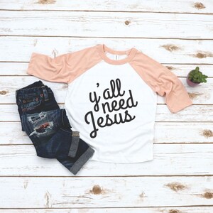 May include: A white and peach colored raglan t-shirt with the text "y'all need Jesus" in black lettering.