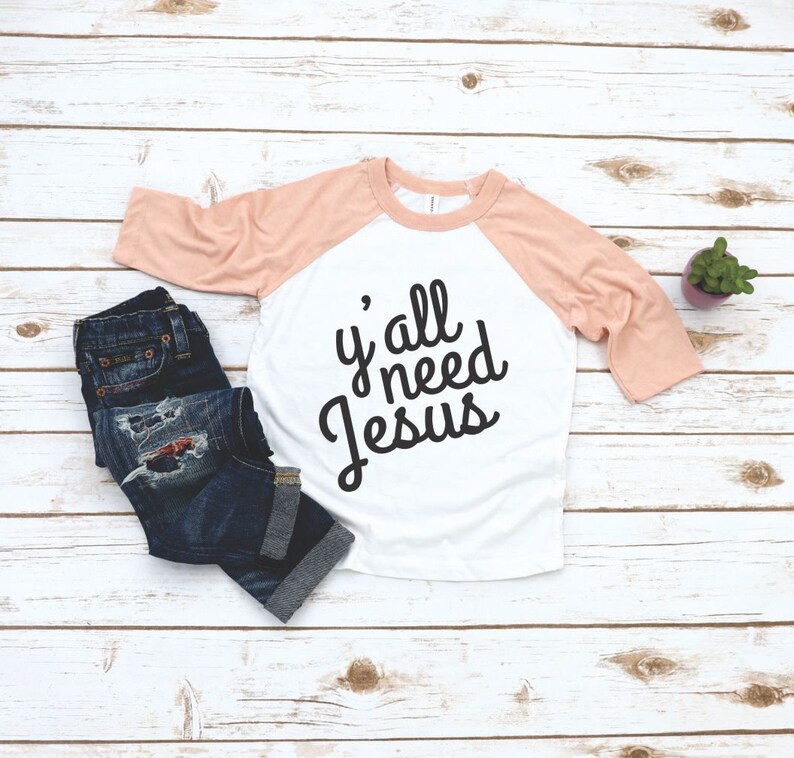 May include: A white and peach colored raglan t-shirt with the text "y'all need Jesus" in black lettering.