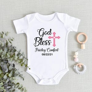 K&ouml;nnte beinhalten: Wei&szlig;er Baby-Body mit dem Text "God Bless" in schwarzer Schrift, einem rosa Kreuz und dem Namen "Paisley Comfort" mit dem Datum "08/22/21". Rechts ist ein Baby-Spielzeug aus Holz und Perlen.