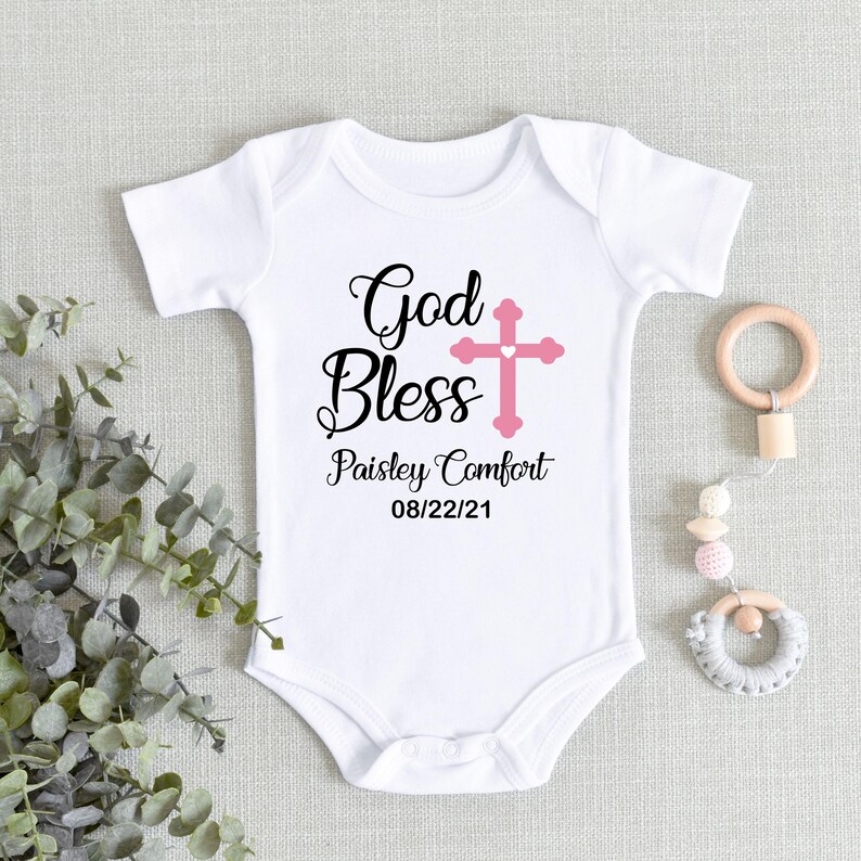 K&ouml;nnte beinhalten: Wei&szlig;er Baby-Body mit dem Text "God Bless" in schwarzer Schrift, einem rosa Kreuz und dem Namen "Paisley Comfort" mit dem Datum "08/22/21". Rechts ist ein Baby-Spielzeug aus Holz und Perlen.