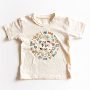 May include: Cream-colored toddler t-shirt with the text "TRUST ME I'M AN ENGINEER" in black. The design features a circular arrangement of colorful engineering-related graphics, including gears, wrenches, and blocks. The shirt is made of soft material.