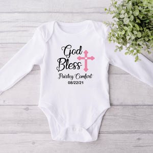 K&ouml;nnte beinhalten: Wei&szlig;er Baby-Body mit langen &Auml;rmeln, mit den Worten "God Bless" in schwarzer Schrift und einem rosa Kreuz. Der Name "Paisley Comfort" und das Datum "08/22/21" sind darunter gedruckt. Der Body liegt auf einer hellen Holzmaserung.