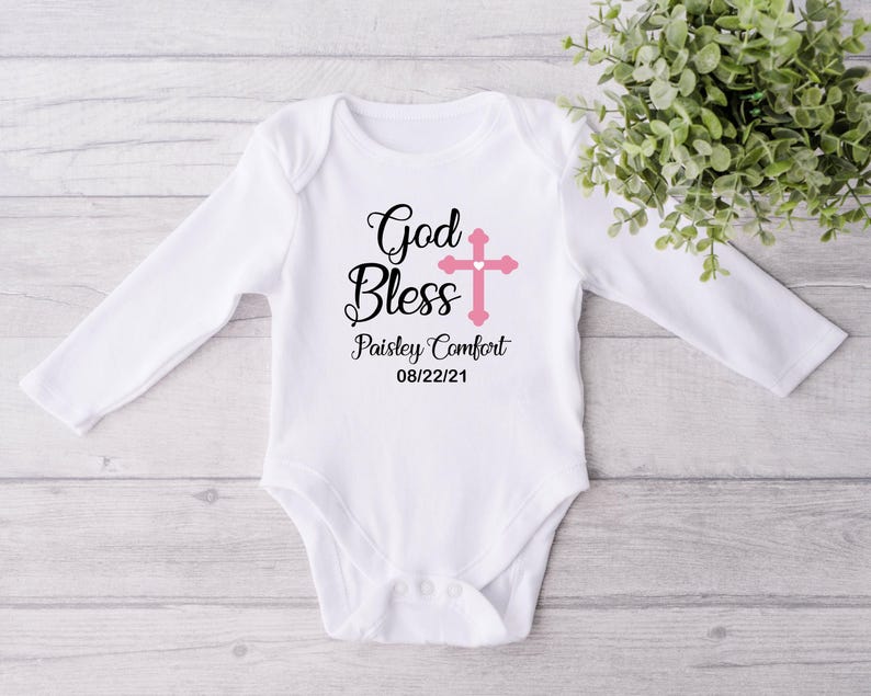 K&ouml;nnte beinhalten: Wei&szlig;er Baby-Body mit langen &Auml;rmeln, mit den Worten "God Bless" in schwarzer Schrift und einem rosa Kreuz. Der Name "Paisley Comfort" und das Datum "08/22/21" sind darunter gedruckt. Der Body liegt auf einer hellen Holzmaserung.
