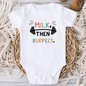 May include: White baby onesie with the text "MILK, THEN BURPEES." in a playful font, with a barbell graphic. The text is in a mix of colors, including orange, green, and black. The onesie is on a woven basket.