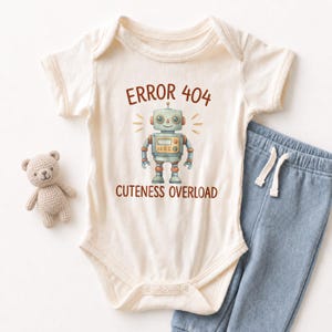 May include: Cream-colored baby onesie with a robot graphic and the text "ERROR 404 CUTENESS OVERLOAD." The onesie is paired with blue pants and a small teddy bear. A cute outfit for a baby.