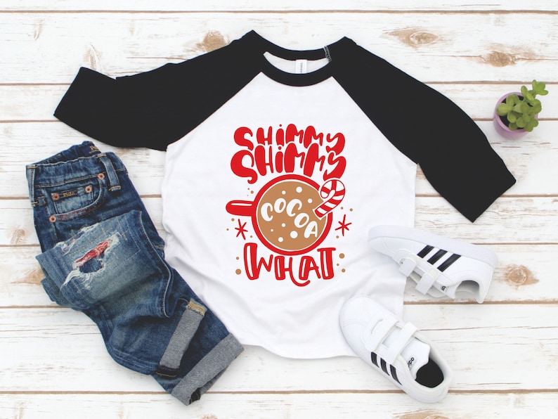 May include: A white and black baseball tee shirt with the text "Shimmy Shimmy Cocoa What" in red lettering. The shirt features a red mug with a brown liquid and a candy cane.
