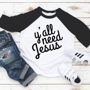 May include: A white and black baseball tee shirt with the text "y'all need Jesus" in black script lettering.