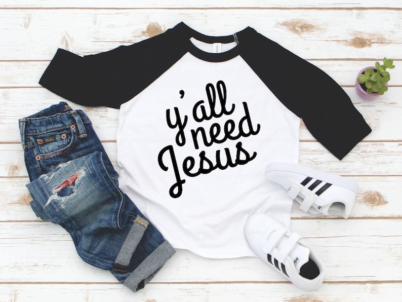 May include: A white and black baseball tee shirt with the text "y'all need Jesus" in black script lettering.