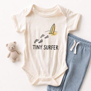 May include: Cream-colored baby bodysuit with the text "TINY SURFER" and a surfboard graphic. The bodysuit is paired with blue pants and a small teddy bear. The graphic includes footprints leading to a surfboard on a beach.