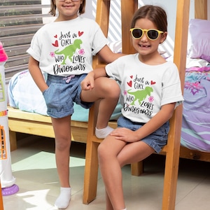 Może przedstawiać: Dwie dziewczynki w białych koszulkach z zielonym dinozaurem i napisem "Just a girl who loves dinosaurs". Dziewczynki noszą okulary przeciwsłoneczne i uśmiechają się.
