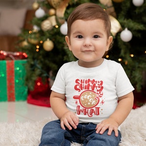 May include: A toddler wearing a white shirt with red text that says "Shimmer Shimmer What" and a picture of a mug with cocoa and a candy cane. The toddler is sitting on a white fluffy rug in front of a Christmas tree.