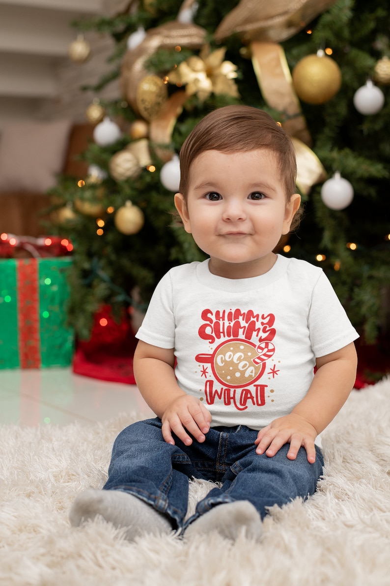 May include: A toddler wearing a white shirt with red text that says "Shimmer Shimmer What" and a picture of a mug with cocoa and a candy cane. The toddler is sitting on a white fluffy rug in front of a Christmas tree.