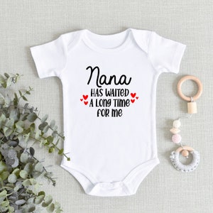 May include: White baby onesie with black text that reads "Nana has waited a long time for me". There are small red hearts around the text.