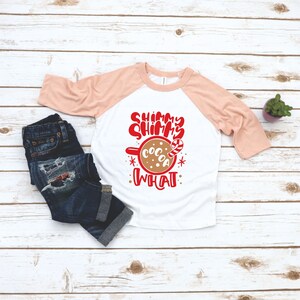 May include: A white and peach colored raglan t-shirt with the text "Shimmy Shimmy Cocoa What" in red. The text is accompanied by a graphic of a mug of hot cocoa with a candy cane.
