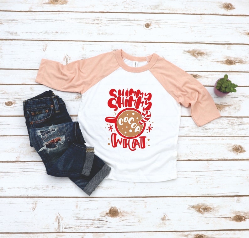 May include: A white and peach colored raglan t-shirt with the text "Shimmy Shimmy Cocoa What" in red. The text is accompanied by a graphic of a mug of hot cocoa with a candy cane.