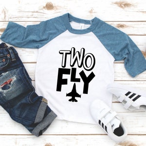 May include: A white and blue baseball tee shirt with the text "TWO FLY" in black with a small airplane graphic. The shirt is on a white wooden surface with a pair of white sneakers with black stripes and a small potted plant.