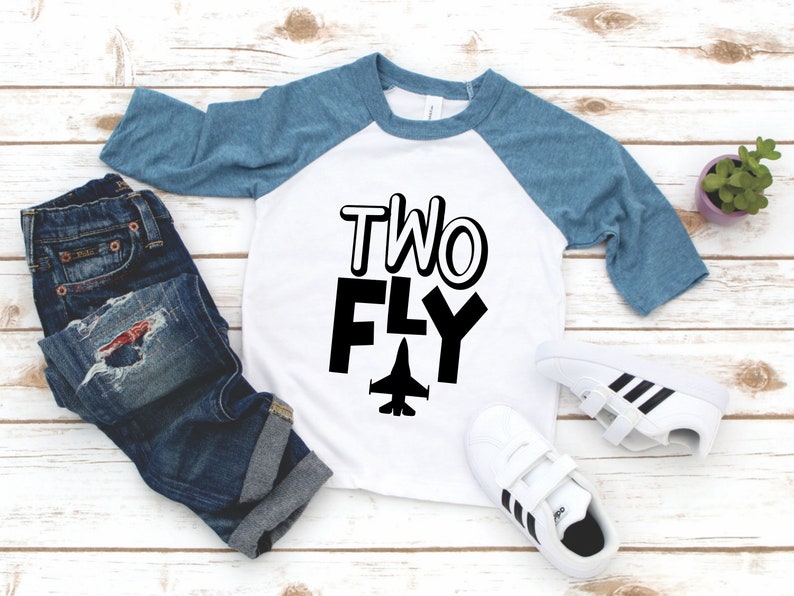 May include: A white and blue baseball tee shirt with the text "TWO FLY" in black with a small airplane graphic. The shirt is on a white wooden surface with a pair of white sneakers with black stripes and a small potted plant.