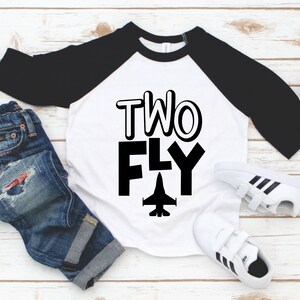 May include: A white and black baseball tee shirt with the text "TWO FLY" and a small airplane graphic. The shirt is a raglan style with black sleeves and a white body.