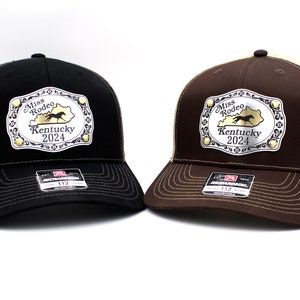 May include: Two trucker hats with a silver buckle featuring a horse silhouette and the text "Miss Rodeo Kentucky 2024". One hat is black with a gray mesh back, and the other is brown with a tan mesh back.