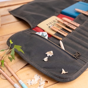 Puede incluir: Un estuche enrollable de lona marrón con cierre de cremallera. El estuche está lleno de lápices y una pequeña libreta que dice "Field Notes".