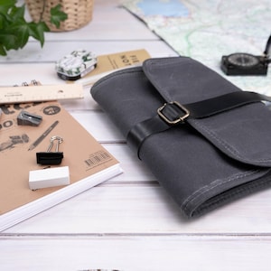 Op de afbeelding: Een grijze canvas etui met een zwarte leren riem en een messing gesp. Een schetsboek, potlood, gum en andere kunstbenodigdheden zijn gerangschikt op een wit houten oppervlak.