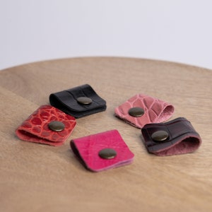 Puede incluir: Seis organizadores de cables de cuero en varios colores, incluyendo negro, rojo, rosa y marrón. Cada organizador tiene un pequeño botón de metal.