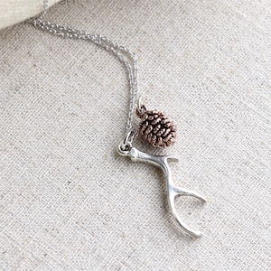 May include: A silver chain necklace with a small antler charm and a pine cone charm. The necklace is lying on a beige fabric surface.