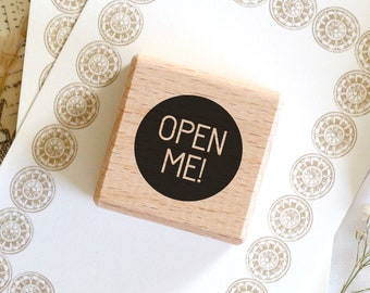 May include: A wooden stamp with the text "OPEN ME!" in a black circle, resting on a white card with a decorative border of circular patterns. A small glass vial with a cork stopper is visible.