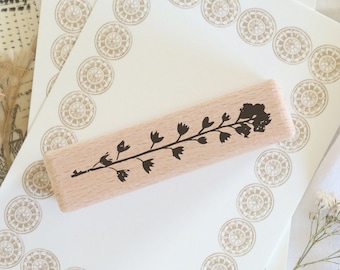 May include: A wooden stamp with a floral design rests on a stack of cream-colored stationery. The stationery is bordered with a repeating circular pattern. Additional stationery and decorative elements are visible in the background.