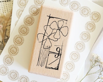 May include: A wooden stamp with a floral design in black ink. The stamp features two flowers, stems, and a rectangular shape with the letter "g". The stamp is on a white surface with decorative circular patterns.