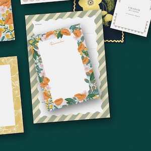 May include: A notepad with a floral design, featuring orange flowers and green leaves around the edge. The word "Remember" is written in cursive. The notepad is on a striped background with other stationery items.