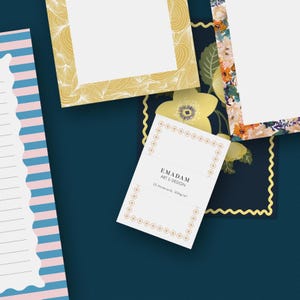 May include: A selection of stationery items on a dark blue surface. The image features patterned frames in gold and floral designs, and a white notecard with the text "EMADAM ART & DESIGN". The notecard is bordered with a gold dotted pattern.