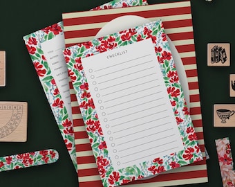 Puede incluir: Un conjunto de artículos de papelería, incluyendo un bloc de notas de lista de verificación con un borde floral de flores rojas y hojas verdes. Debajo hay un cuaderno a rayas rojas y blancas. Sellos de madera y cinta washi floral también están presentes.